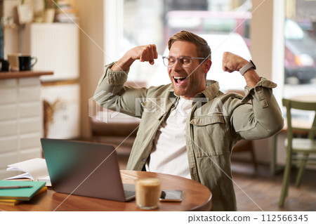 Portrait of happy young man in glasses rejoicing, sitting in cafe, watching sports game on laptop, celebrating, showing big muscles and laughing Portrait of happy young man in glasses rejoicing, sitting in cafe, watching sports game on laptop, celebrating, showing big muscles and laughing 112566345