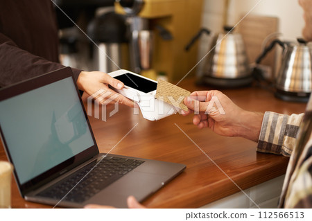 Cropped shot of male hand paying contactless for his order in coffee shop, sitting at counter with laptop, pressing card to POS terminal, working from home Cropped shot of male hand paying contactless for his order in coffee shop, sitting at counter with laptop, pressing card to POS terminal, working from home 112566513