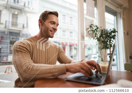 Portrait of handsome young man sitting in cafe with laptop. Digital nomad, freelancer working outdoors from coffee shop, typing something Portrait of handsome young man sitting in cafe with laptop. Digital nomad, freelancer working outdoors from coffee shop, typing something 112566610