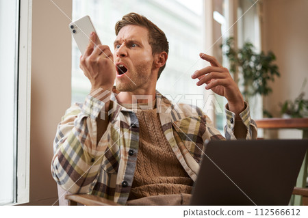 Portrait of an angry man shouting at his mobile phone. Frustrated cafe visitor arguing with someone on call, sitting with laptop near window, screaming at telephone Portrait of an angry man shouting at his mobile phone. Frustrated cafe visitor arguing with someone on call, sitting with laptop near window, screaming at telephone 112566612