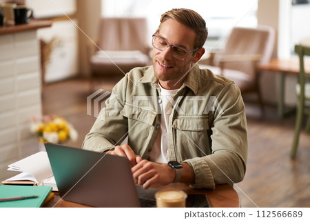 Remote work and freelance concept. Handsome man in glasses sits with notebook and laptop, studying online, working remotely from coffee shop, sitting in cafe with computer Remote work and freelance concept. Handsome man in glasses sits with notebook and laptop, studying online, working remotely from coffee shop, sitting in cafe with computer 112566689