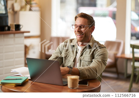 Image of young digital nomad, man in glasses sits in cafe, works from coffee shop, uses laptop in co-working space, wears glasses, drinks his beverage Image of young digital nomad, man in glasses sits in cafe, works from coffee shop, uses laptop in co-working space, wears glasses, drinks his beverage 112566716