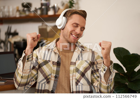 Portrait of handsome happy guy, freelancer working and listening to music in wireless headphones, dancing on his chair in cafe, enjoying favourite song in earphones Portrait of handsome happy guy, freelancer working and listening to music in wireless headphones, dancing on his chair in cafe, enjoying favourite song in earphones 112566749