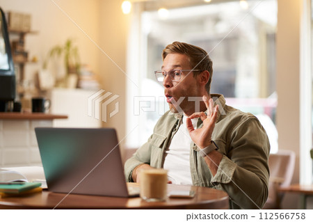 Portrait of young freelancer, digital nomad having an online meeting, video chats, shows okay sign at laptop, confirm something, makes ok hand gesture, sits in cafe, works on remote 112566758