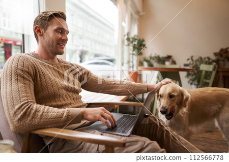 Handsome young man working in cafe with a dog, sitting on chair and using laptop, petting his golden retriever in animal-friendly co-working space Handsome young man working in cafe with a dog, sitting on chair and using laptop, petting his golden retriever in animal-friendly co-working space 112566778