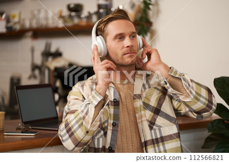 Portrait of handsome happy guy, freelancer working and listening to music in wireless headphones, dancing on his chair in cafe, enjoying favourite song in earphones Portrait of handsome happy guy, freelancer working and listening to music in wireless headphones, dancing on his chair in cafe, enjoying favourite song in earphones 112566821