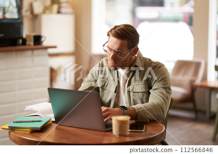 Portrait of businessman in glasses, sits in cafe with laptop, looks concentrated at screen with his project task, works remotely from coffee shop Portrait of businessman in glasses, sits in cafe with laptop, looks concentrated at screen with his project task, works remotely from coffee shop 112566846
