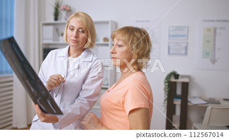 A concentrated traumatologist and a senior patient examine an X-ray image, discussing the diagnosis 112567014