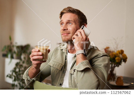 Image of smiling, good-looking young blond man with phone, drinking coffee in cafe, talking to someone over the telephone, answering a call Image of smiling, good-looking young blond man with phone, drinking coffee in cafe, talking to someone over the telephone, answering a call 112567062