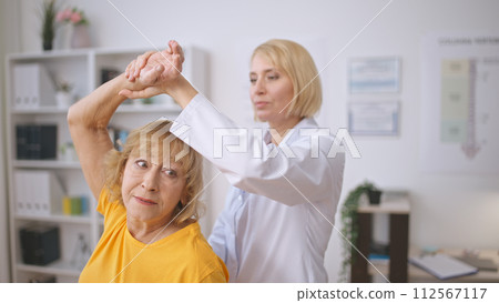 A masseur assists a senior lady with muscle stretching, providing treatment at a rehabilitation center A masseur assists a senior lady with muscle stretching, providing treatment at a rehabilitation center 112567117