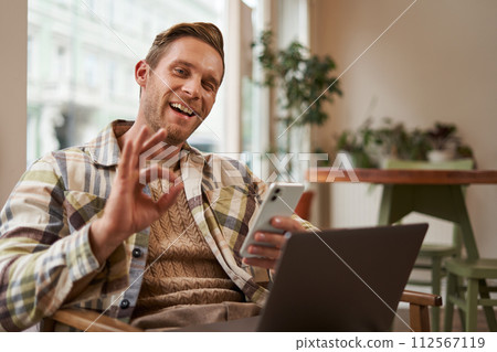 Portrait of handsome man in cafe, sitting with laptop, working remotely, freelancer with phone in hands, shows okay, ok gesture in approval, agree with something, like product 112567119
