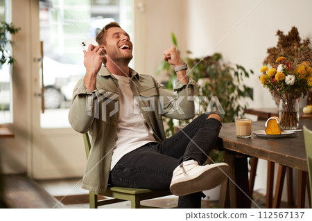 Portrait of relieved, happy young man, listens to happy news or podcast in earphones, sits in coffee shop, holds smartphone, raises hands up and smiling from excitement, spending time in cafe 112567137