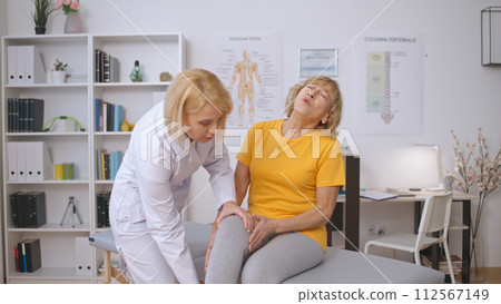 A woman traumatologist examines the sore knee of a female patient who is crying in pain A woman traumatologist examines the sore knee of a female patient who is crying in pain 112567149