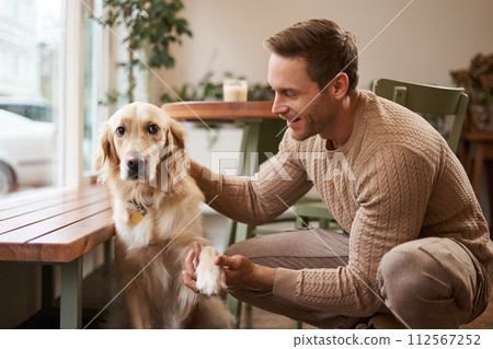 Portrait of young handsome man with his dog, sitting in cafe, golden retriever gives paw and looks at his owner Portrait of young handsome man with his dog, sitting in cafe, golden retriever gives paw and looks at his owner 112567252