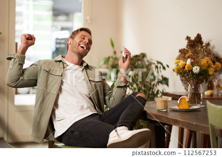 Portrait of handsome happy man in cafe, listens to music in wireless earphones, holding smartphone, connects to public wifi and enjoys favourite song, relaxing in coffee shop Portrait of handsome happy man in cafe, listens to music in wireless earphones, holding smartphone, connects to public wifi and enjoys favourite song, relaxing in coffee shop 112567253