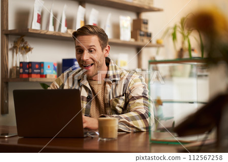 Portrait of handsome young digital nomad, man working in cafe on laptop, looking happy and pleased with his online project, sitting in co-working coffee shop 112567258