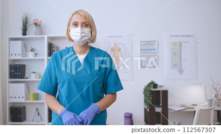 A woman doctor in a face mask dons medical gloves, preparing for a check-up or procedure A woman doctor in a face mask dons medical gloves, preparing for a check-up or procedure 112567307