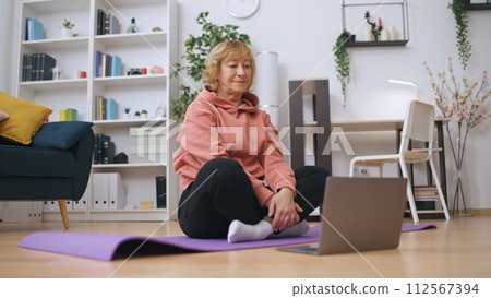 A senior woman communicates with a fitness trainer online, exercising at home 112567394