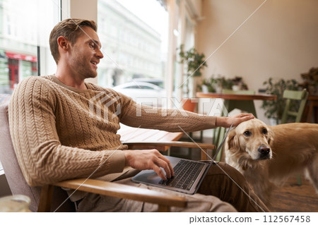 Portrait of young man, digital nomad, sitting in cafe near window, using laptop, working with his pet, petting golden retriever while sitting in chair with computer on laps Portrait of young man, digital nomad, sitting in cafe near window, using laptop, working with his pet, petting golden retriever while sitting in chair with computer on laps 112567458