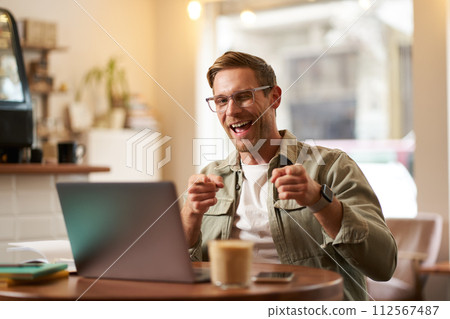 Happy young man in glasses, freelancer sitting in cafe with laptop, drinking coffee, pointing fingers at camera and smiling cheerfully 112567487