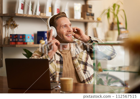 Lifestyle portrait of smiling digital nomad, man in cafe, working on laptop, listening music in wireless headphones and smiling, dancing on chair while enjoying favourite song Lifestyle portrait of smiling digital nomad, man in cafe, working on laptop, listening music in wireless headphones and smiling, dancing on chair while enjoying favourite song 112567515
