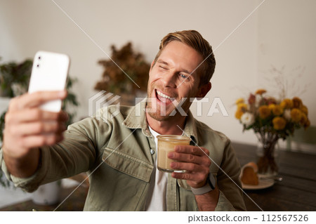 Close up portrait of happy, charismatic young blond man, taking selfie on smartphone, posing with cup of coffee, enjoys his drink in cafe, recommending a place to his followers online 112567526