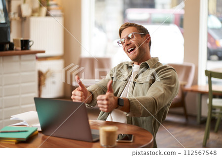 Portrait of satisfied, happy young man in glasses, working in cafe, sitting in co-working space with laptop, showing thumbs up, like and approve smth good, chatting, giving online lessons 112567548