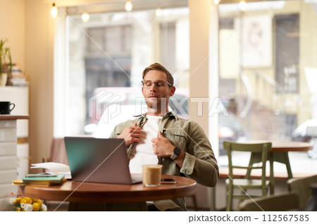 Portrait of businessman in glasses, sits in cafe with laptop, looks concentrated at screen with his project task, works remotely from coffee shop Portrait of businessman in glasses, sits in cafe with laptop, looks concentrated at screen with his project task, works remotely from coffee shop 112567585