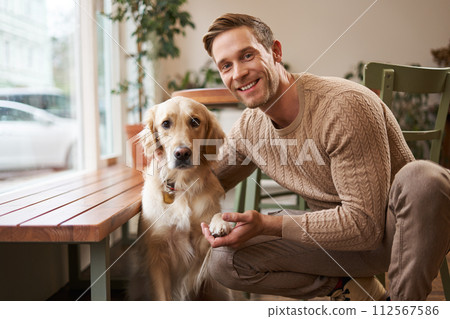 Portrait of a lovely golden retriever and handsome man in cafe. Cute dog gives paw and takes picture with his owner in coffee shop Portrait of a lovely golden retriever and handsome man in cafe. Cute dog gives paw and takes picture with his owner in coffee shop 112567586