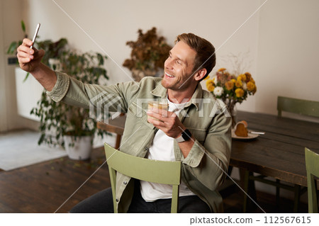 Portrait of handsome happy young man, taking selfie in cafe, posing with cup of coffee, video chats using smartphone app, talks to a friend 112567615