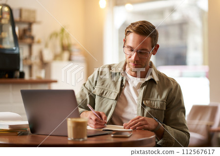 Portrait of handsome young guy in glasses, man studying, taking notes, looking at laptop screen during online lesson, attends a meeting and writing down information, sitting in cafe 112567617