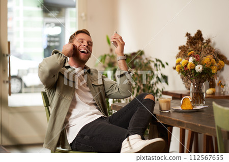 Lifestyle portrait of handsome young man, sitting in cafe, talking to a friend on mobile phone with wireless earphones, laughing and smiling, listening to funny podcast Lifestyle portrait of handsome young man, sitting in cafe, talking to a friend on mobile phone with wireless earphones, laughing and smiling, listening to funny podcast 112567655