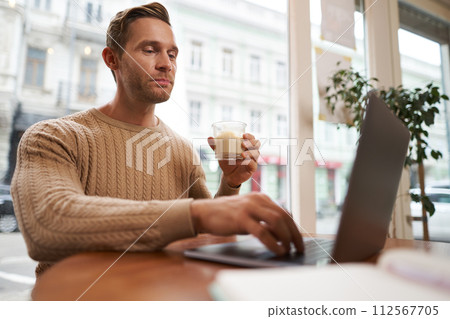Portrait of handsome young man sitting in cafe with laptop. Digital nomad, freelancer working outdoors from coffee shop, typing something Portrait of handsome young man sitting in cafe with laptop. Digital nomad, freelancer working outdoors from coffee shop, typing something 112567705