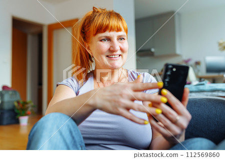 portrait red-haired woman using mobile phone while sitting on sofa at home in living room 112569860