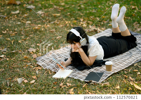 Young woman with headphone relaxing on green lawn in the city park and using mobile phone 112570821