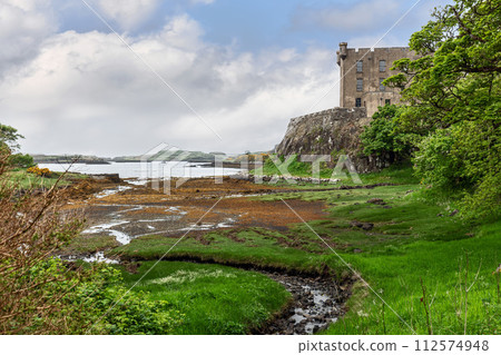 Historic Dunvegan Castle on Isle of Skye, surrounded by vibrant green flora under a dynamic sky 112574948