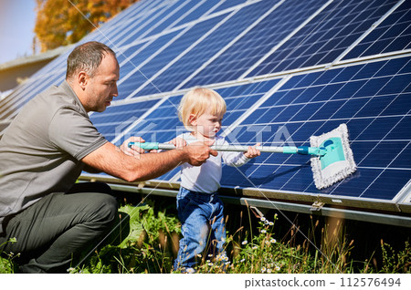 Father showing his little son the solar panels during sunny day. Cute boy helping his dad to clean solar battery. Young man teaching his small kid how to care about their source of energy. 112576494