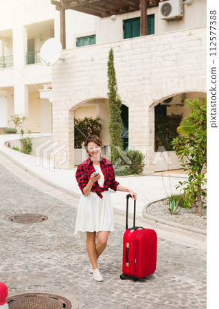 Young woman in a red shirt with a suitcase and phone on the city street.. 112577388