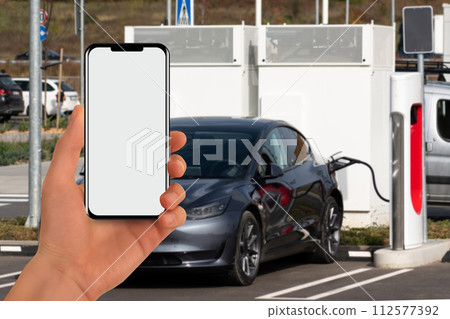 A hand with a phone on the background of a charging station for electric cars 112577392