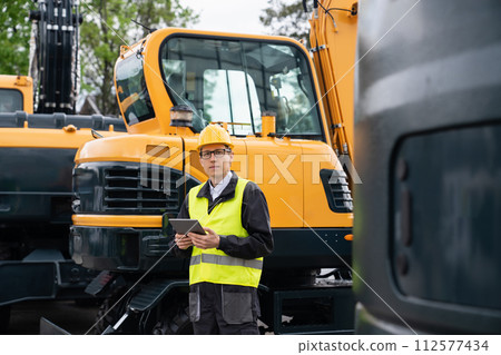 Engineer in a helmet with a digital tablet stands next to construction excavators.. 112577434