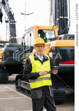 Engineer in a helmet with a digital tablet stands next to construction excavators.. 112577440