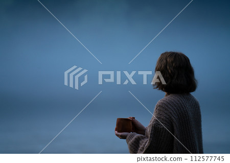 A woman in a sweater on the bank of a foggy river in the evening.. A woman in a sweater on the bank of a foggy river in the evening.. 112577745