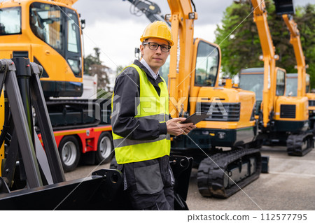 Engineer in a helmet with a digital tablet stands next to construction excavators.. 112577795