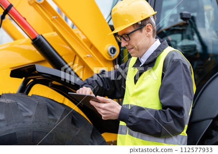 Engineer in a helmet with a digital tablet stands next to construction excavators.. 112577933