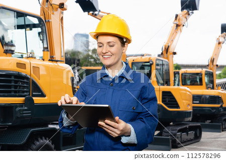 Engineer in a helmet with a digital tablet stands next to construction excavators. 112578296
