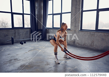 Focused young athlete woman working out using battle ropes with deep squat in urban fitness space. Motivation. Focused young athlete woman working out using battle ropes with deep squat in urban fitness space. Motivation. 112579440