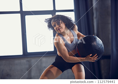 Dynamic portrait of young athletic curly woman in sportswear doing exercise with medicine ball in motion as strength training in gym. Dynamic portrait of young athletic curly woman in sportswear doing exercise with medicine ball in motion as strength training in gym. 112579441