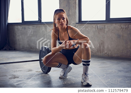 Young, beautiful woman with athletic body sitting in squat next to barbell and resting between trainings in gym. Young, beautiful woman with athletic body sitting in squat next to barbell and resting between trainings in gym. 112579479