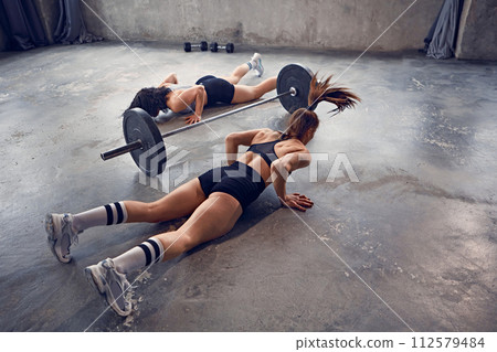 Dynamic portrait of young women dressed sportswear warming up doing some push ups in motion near barbell at gym. 112579484