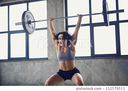 Side view of focused young strong woman in sportswear doing squat technique with barbell overhead in motion at gym. Side view of focused young strong woman in sportswear doing squat technique with barbell overhead in motion at gym. 112579511
